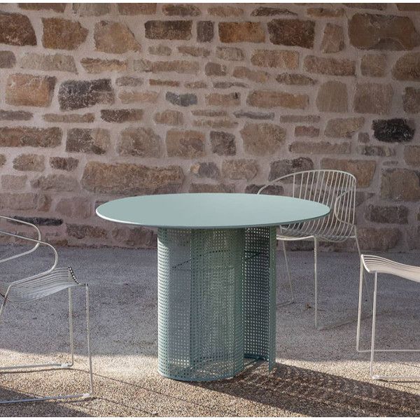 Round metal Arena dining table with a pedestal base, set against a stone wall with wire chairs.