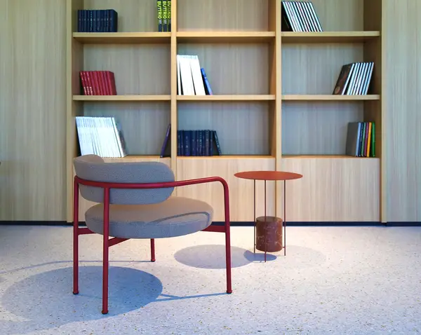 Giotto lounge chair with grey upholstery and a crimson frame positioned near a small matching table by a wood bookcase.