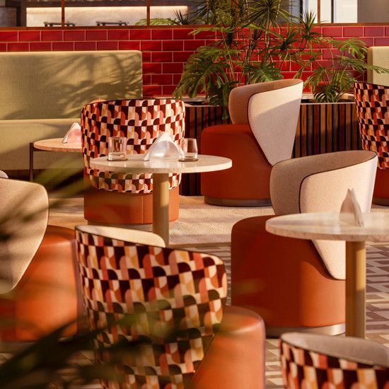 Yana pouf with beige upholstery and wooden frame, set in a lounge with patterned chairs and red brick.