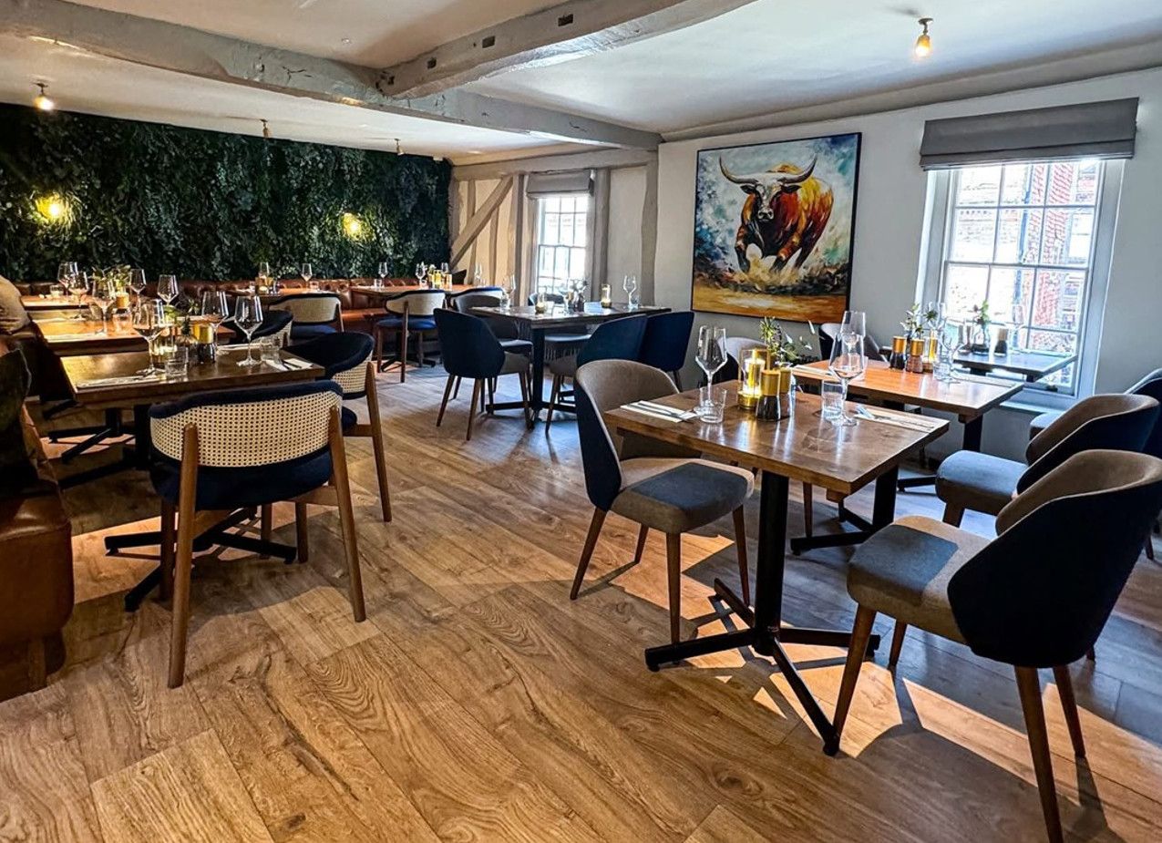 Heavens Kitchen Dorking dining area. Tables set with glasses, chairs, wood floor, and bull painting.