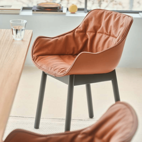 Side view of a Baltic Soft W Armchair in tan upholstery with black legs, next to a table and a glass of water.