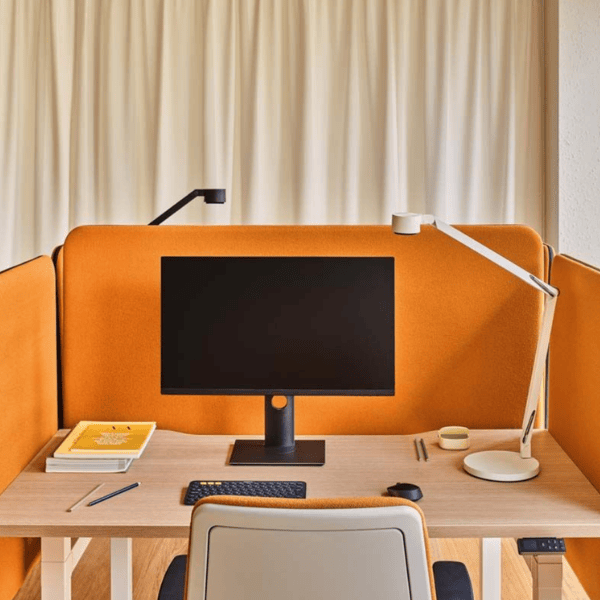 Angled view of Sonic Sit Stand Desk Screen in orange fabric, showing wood frame, acoustic filling, and desk setup with monitor.