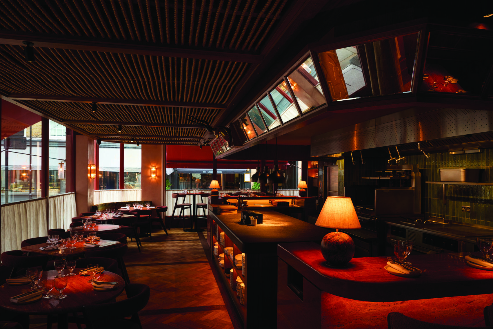 kudu restaurant Kudu Marylebone restaurant interior with warm lighting, wooden ceiling, tables, and a view of the kitchen area.