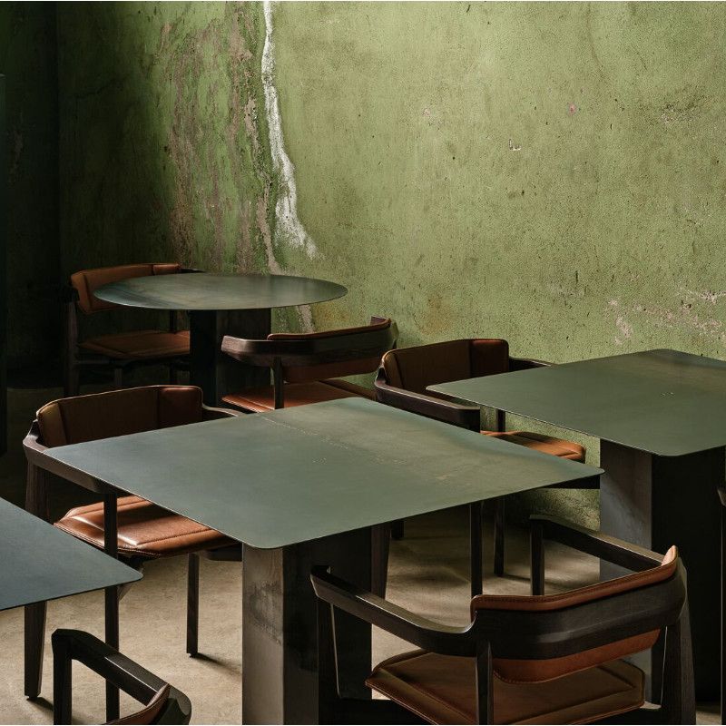 Rasa Armchair side view. Upholstered seat with a wooden frame, in a lounge setting against a textured green wall.