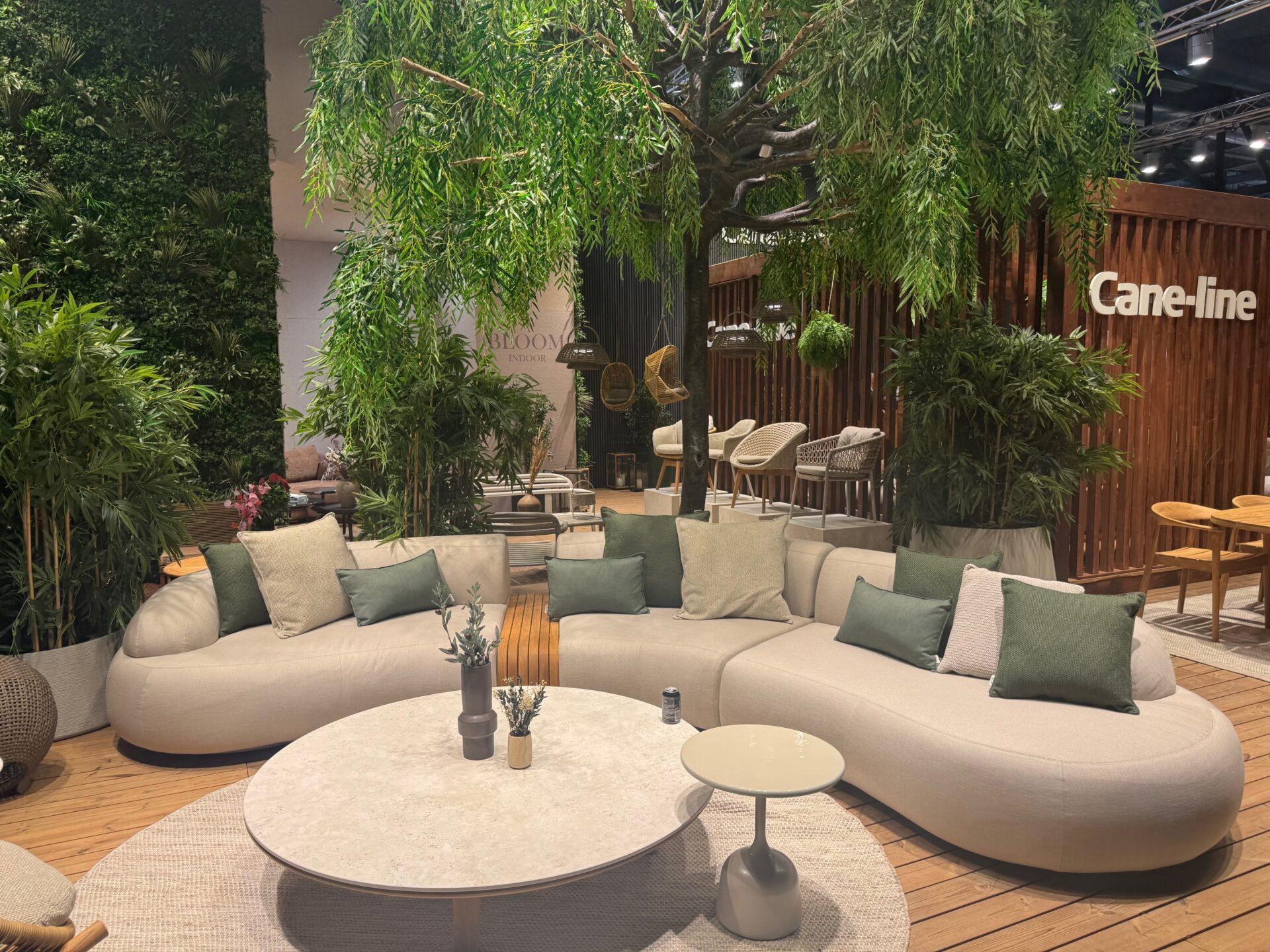 Salone del Mobile 2026 hospitality trend: Cane-line modular sofa with green cushions, surrounded by lush greenery and natural elements.