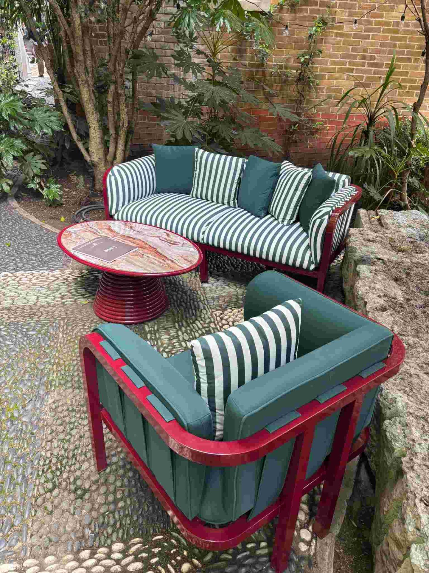Outdoor seating area displays a striped sofa, matching armchair and marble-topped table with red frames on a stone patio, contract outdoor furniture.