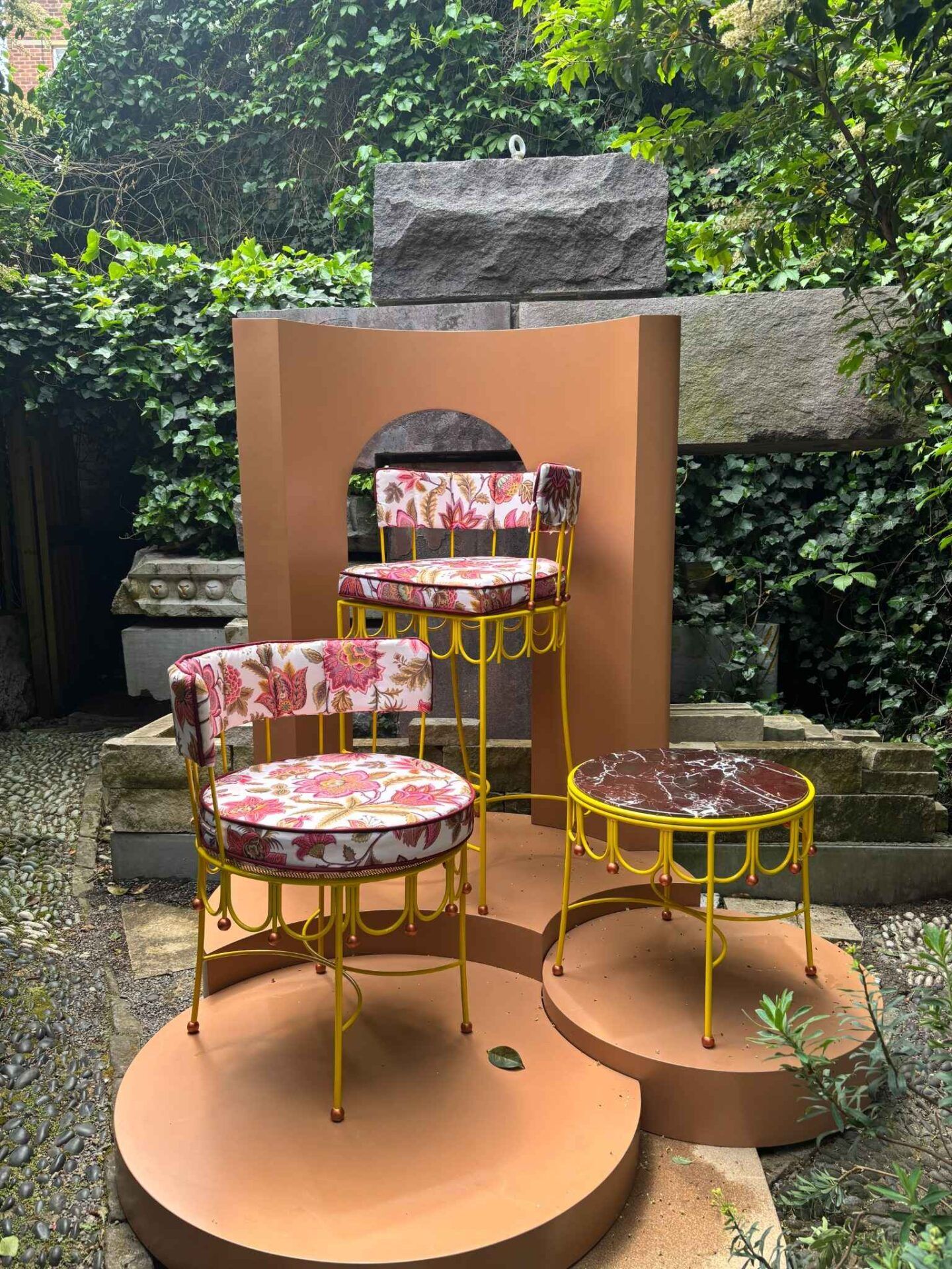 Yellow metal framed chairs and a table with floral upholstery are displayed outdoors on a terra cotta platform.