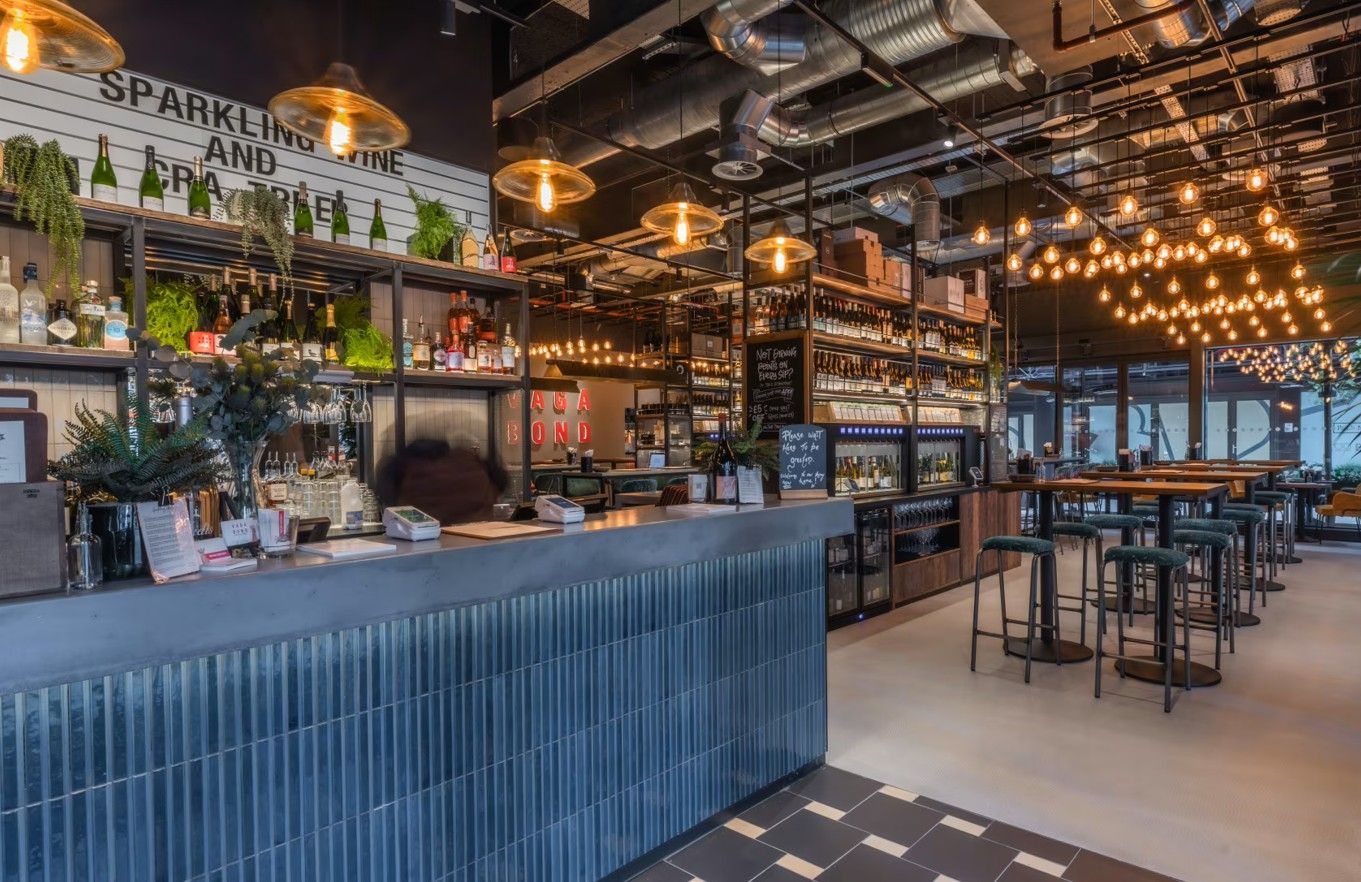 Vagabond Broadgate's bar area features a blue corrugated counter, shelves stocked with bottles, and an array of glowing pendant lights, contract furniture.