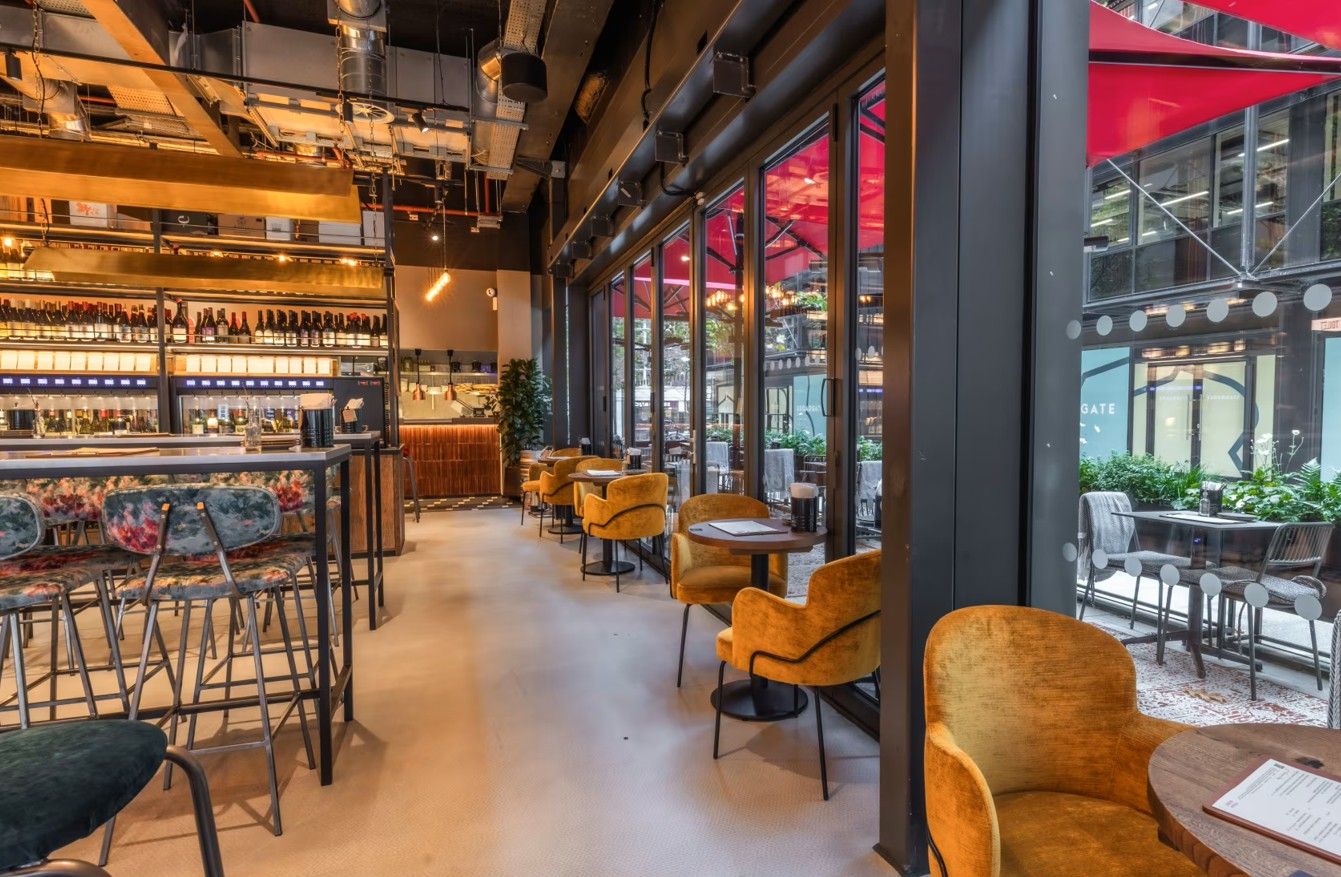 Vagabond Broadgate11 Within a bar area, the round Caldas Coffee Table with a dark metal base is pictured next to comfortable mid-century modern style chairs.