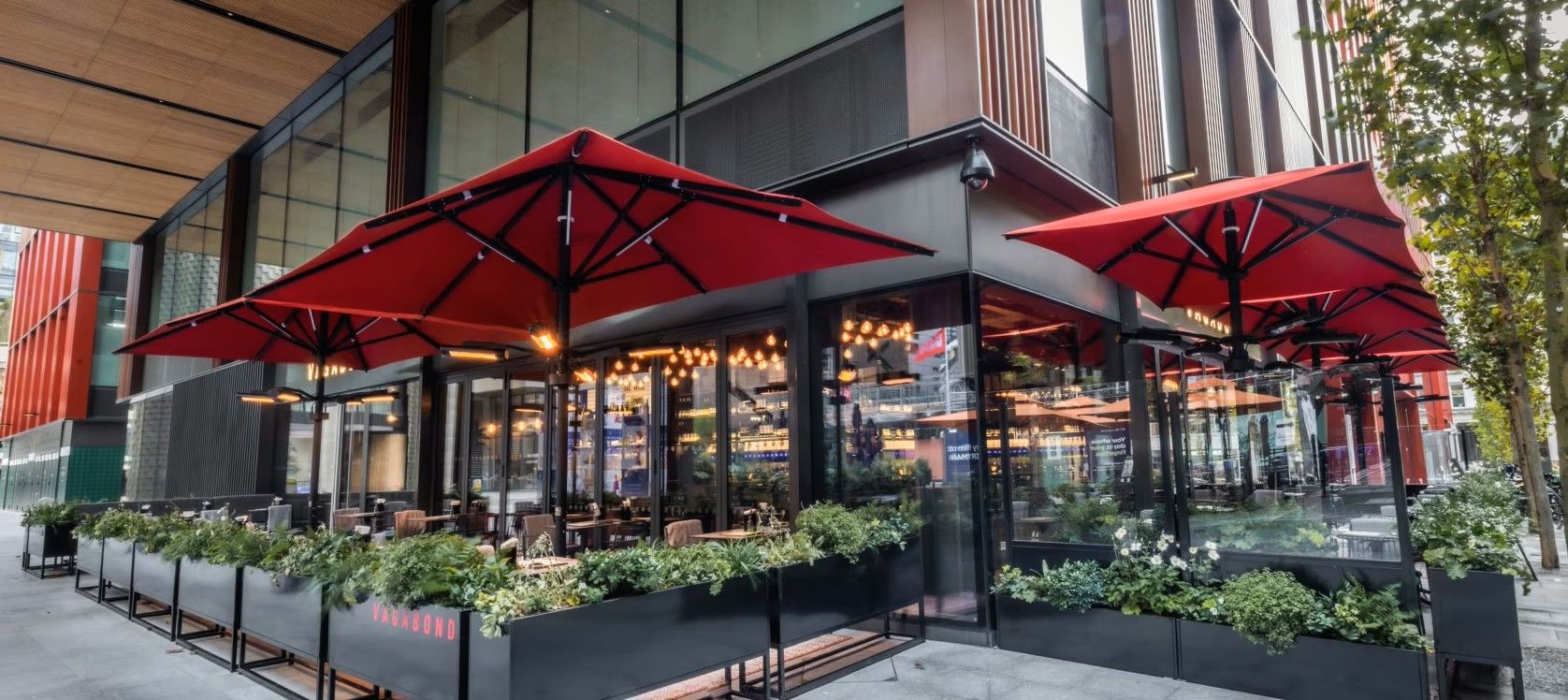 Vagabond Broadgate12 Captured is an outdoor restaurant seating area, featuring Caldas Coffee Tables beneath vibrant red umbrellas, perfect for commercial or outdoor use.