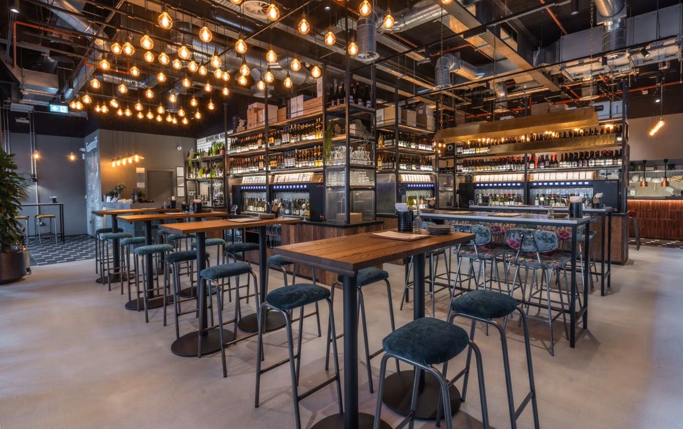 Vagabond Broadgate2 Inside Vagabond Broadgate, wooden tables paired with blue upholstered stools sit before shelves displaying wine bottles and an open bar.