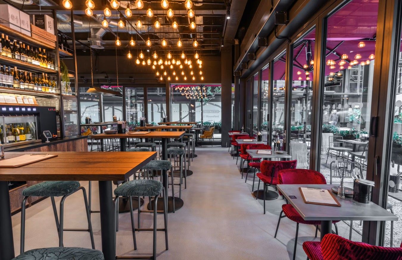 Vagabond Broadgate3 Within a warmly illuminated restaurant, a Caldas Coffee Table with a light gray top is shown among the many tables and seating areas.