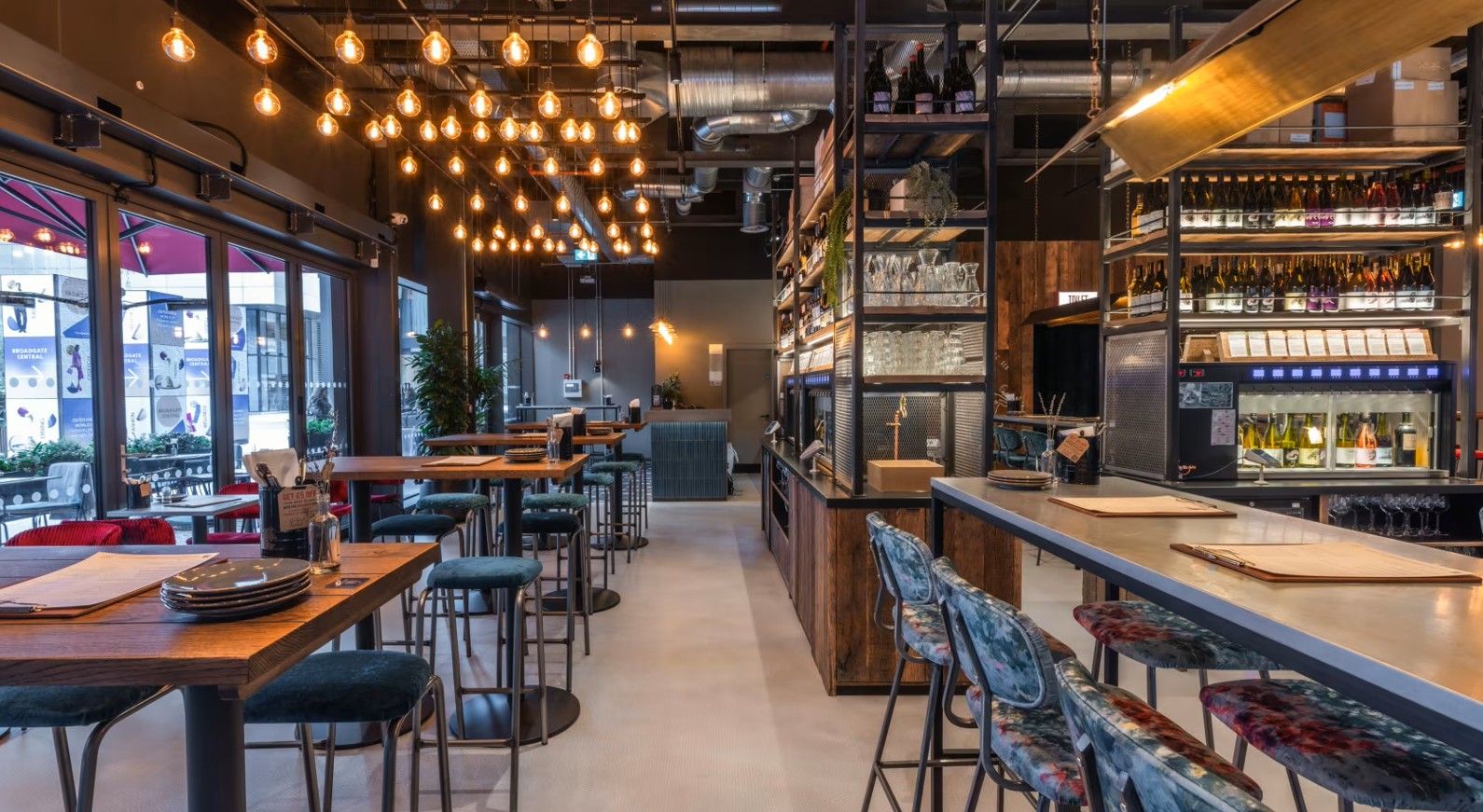Vagabond Broadgate5 Pictured within a restaurant, a Caldas Coffee Table with a wood top and metal base is shown, positioned near comfortable bar seating.