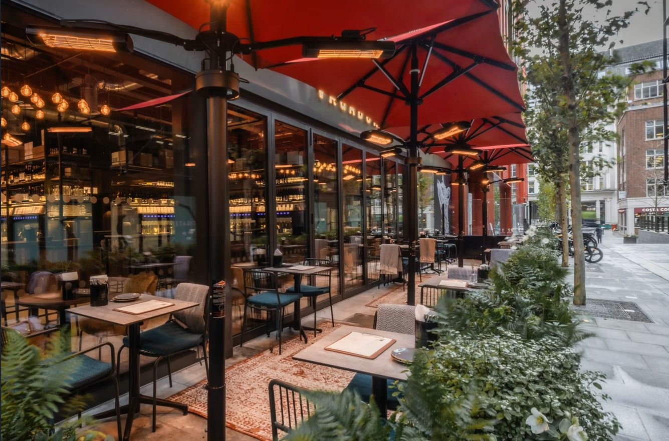 Vagabond Broadgate6 From an offset perspective, the Caldas Coffee Table with a square top and metal base sits on a rug in an outdoor restaurant setting.