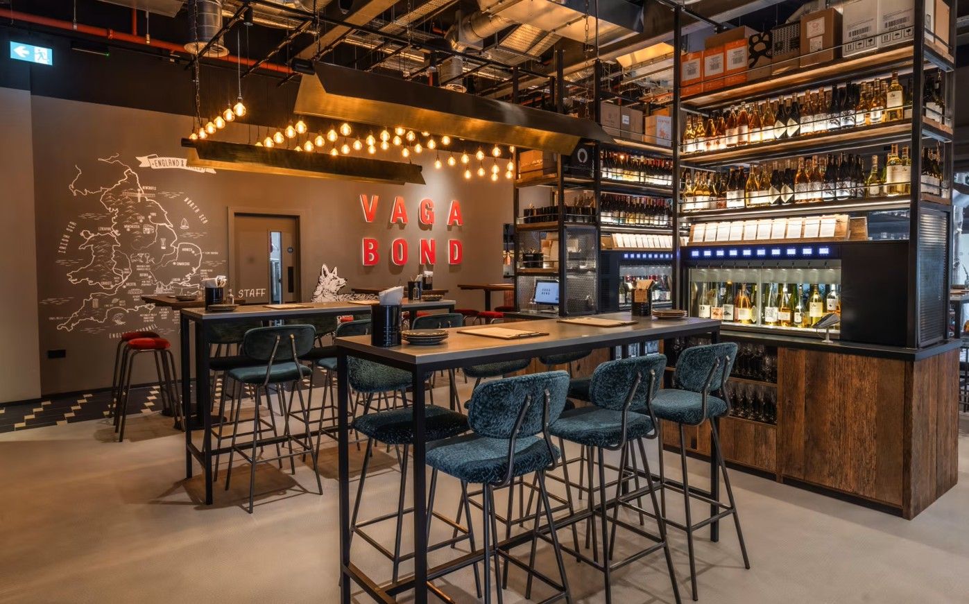 Vagabond Broadgate7 Within a modern bar, a Caldas Coffee Table setup features a metal frame and dark surface, complemented by plush teal barstools, and beverage shelving.