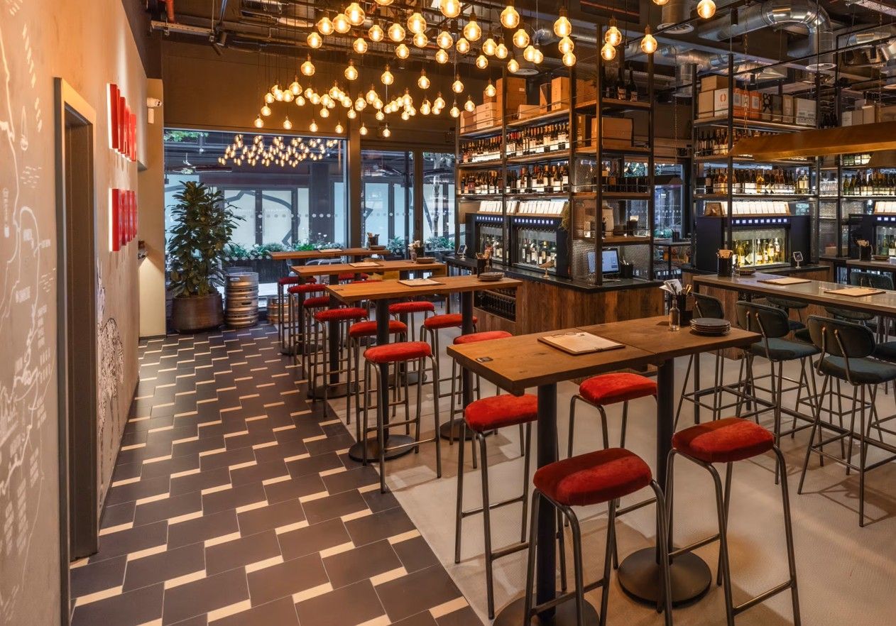 Vagabond Broadgate9 A look inside a bar showcasing several Caldas Coffee Tables with wooden tops, paired with red barstools, under a canopy of lights.