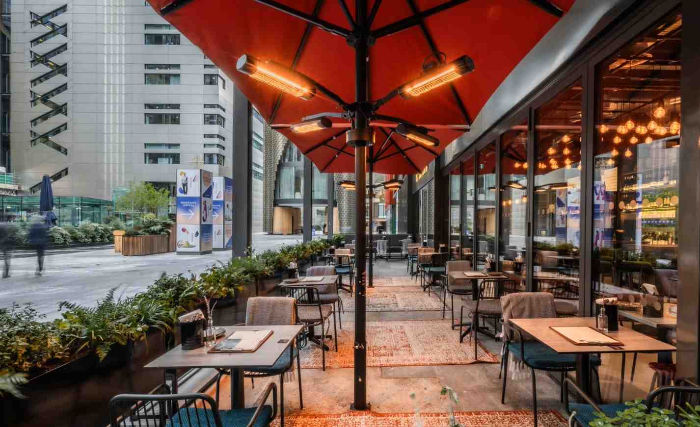 vagabond_broadgate14_100kb Patio at Vagabond Broadgate shows tables with blue chairs under red umbrellas with heaters, and rugs on the concrete floor.