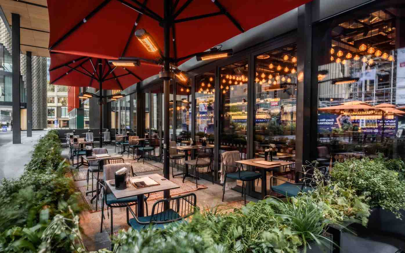 vagabond_outdoor_100kb Vagabond Broadgate's outdoor seating area features red umbrellas, heaters, tables, chairs, and potted plants.