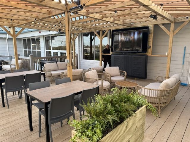 Bashley Holiday Park Large flat-screen television mounted on a wooden structure above a patio seating area with dining tables and armchairs.
