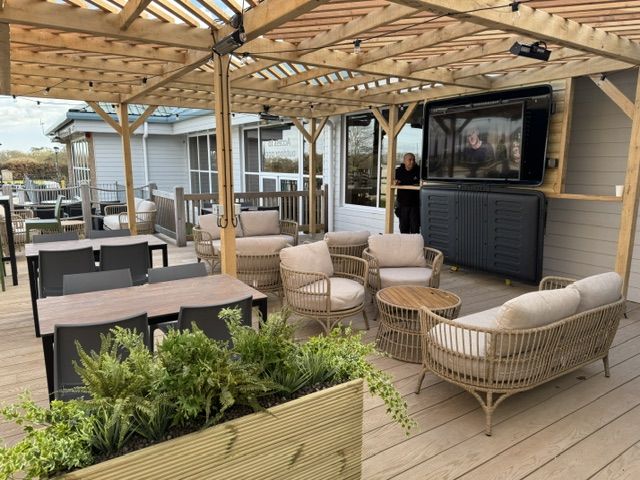 Bashley Holiday Park 3 Potted green ferns sit in the foreground of a sunlit deck featuring woven lounge furniture and a wall-mounted TV.