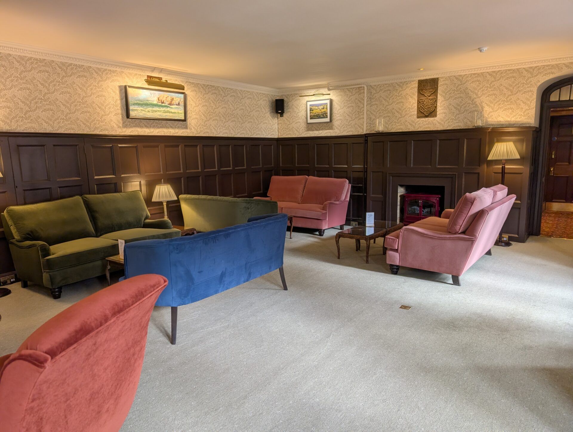 PXL_20260321_125323135 Velvet sofas in blue, pink, and olive green occupy a wood-paneled hotel lounge with light-patterned wallpaper.
