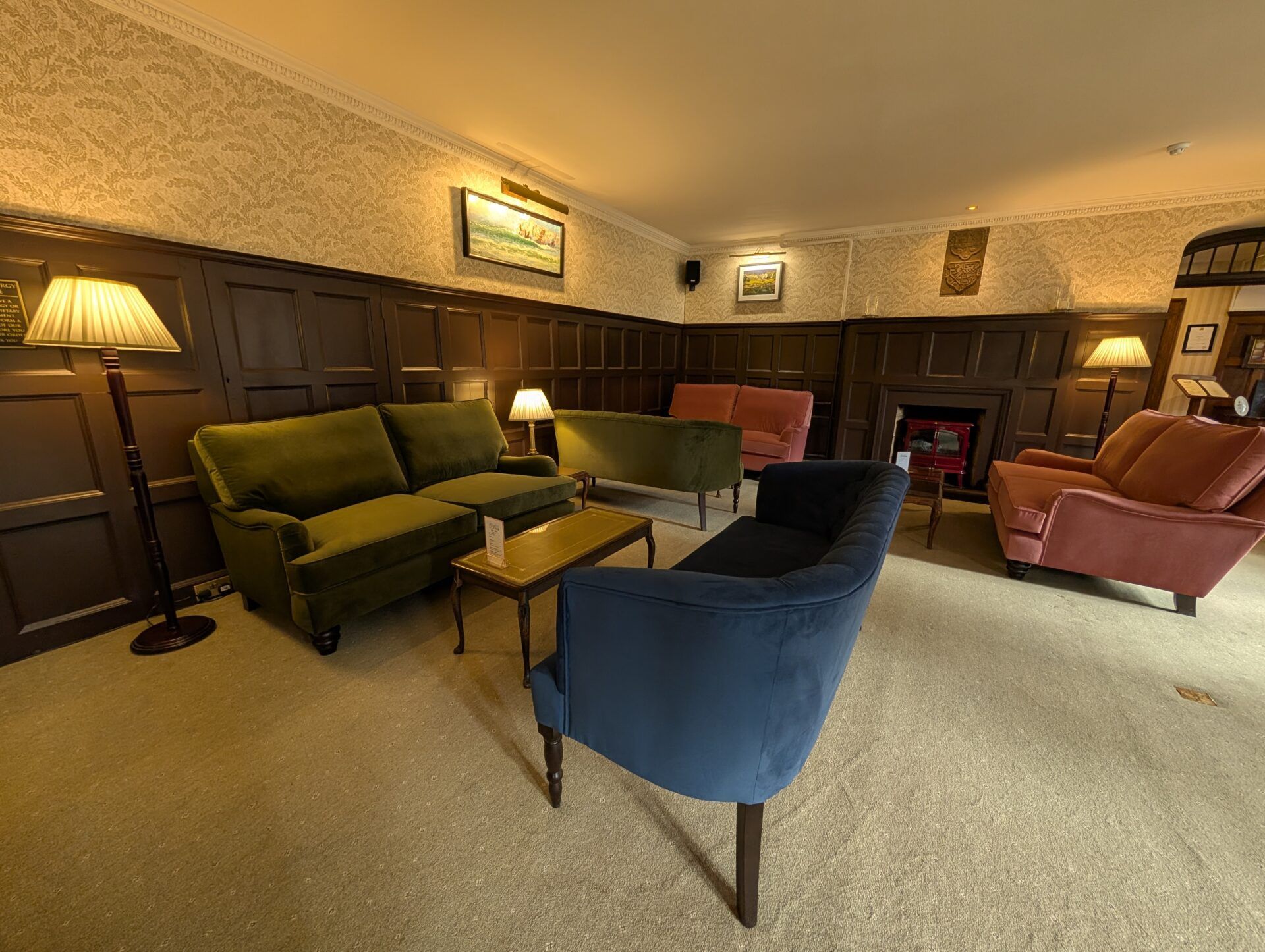 PXL_20260321_125334006 Blue curved sofa anchors a hotel lounge furnished with green, pink, and terracotta seating against dark wood paneling.