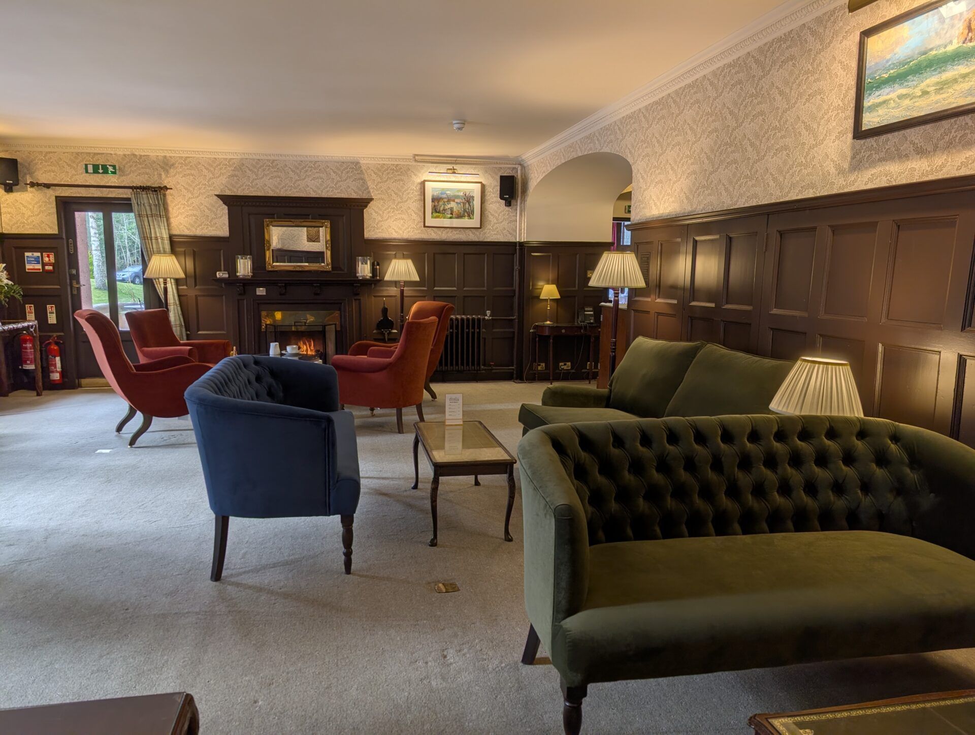 PXL_20260321_125355548 Tufted olive green sofa occupies the foreground of a wood-paneled hotel lounge with red chairs near a lit fireplace.