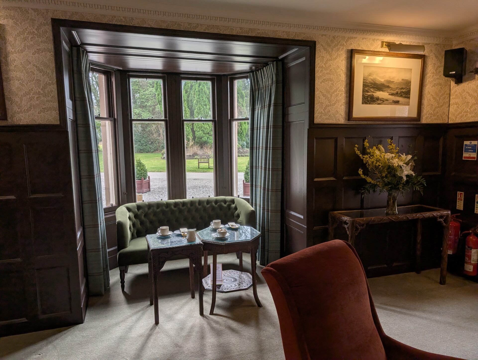 PXL_20260321_125425992 Olive green tufted bench sits below a bay window overlooking a garden, paired with two small tables set for tea.
