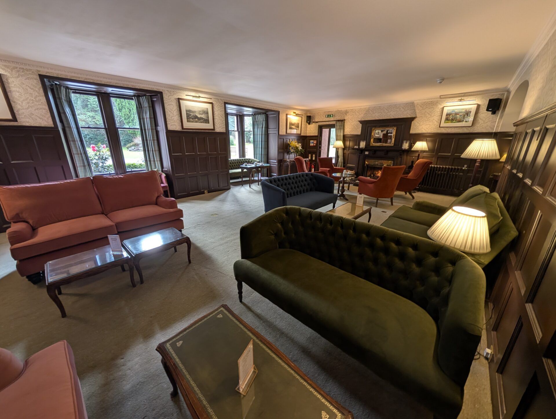 PXL_20260321_125446915 Wide lounge features a dark green velvet chesterfield sofa, a terracotta couch, and vibrant red chairs near a fireplace.