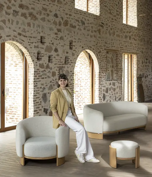 Woman sitting on the stylish cream Hug Lounge Chair in a spacious room with rustic stone walls and archways.