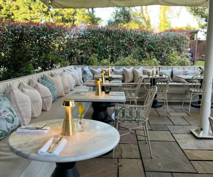 L-shaped cushioned bench seating with patterned pillows lines a sunlit patio featuring marble tables and brass lamps.