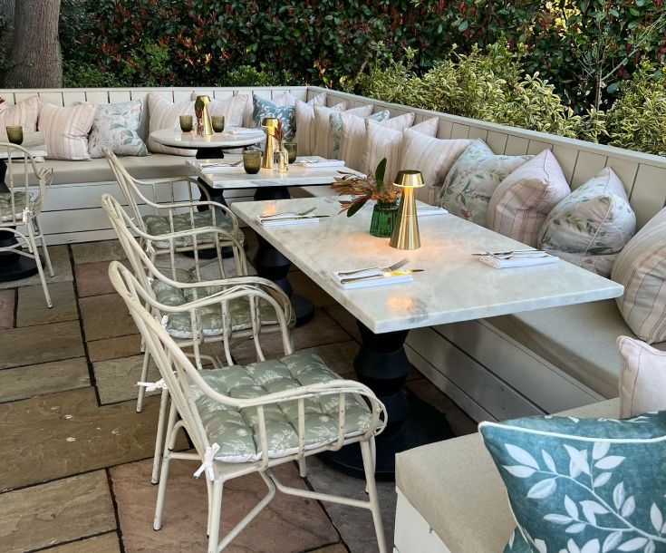 the victoria4 Illuminated brass lamps cast a warm glow over marble tables arranged with cutlery in a leafy outdoor seating terrace.