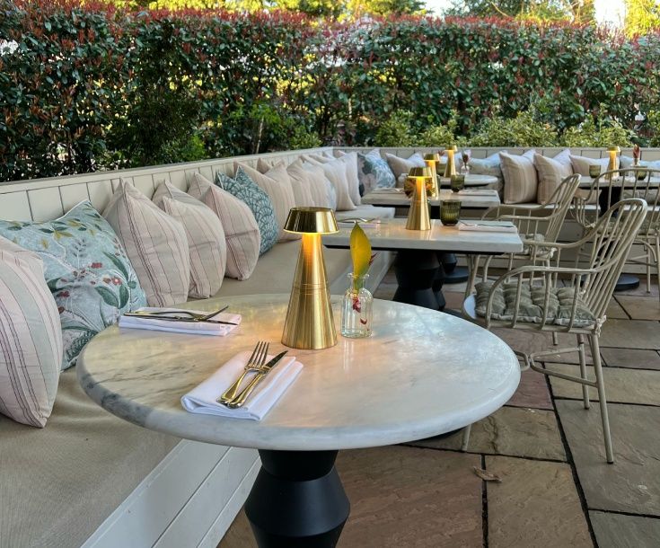 the victoria6 Round marble bistro tables with golden conical lamps and white napkins sit beside a long bench lined with plush pillows.