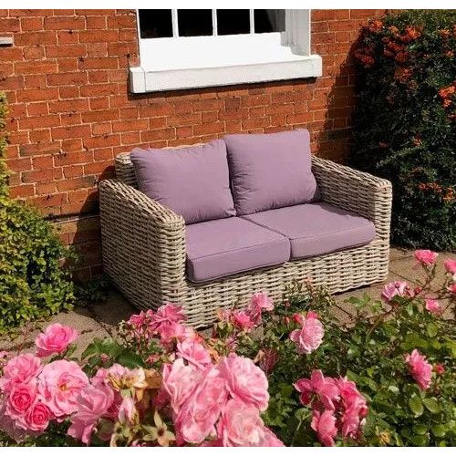 Tula sofa with woven frame and mauve cushions placed on a sunny garden patio beside a red brick wall.