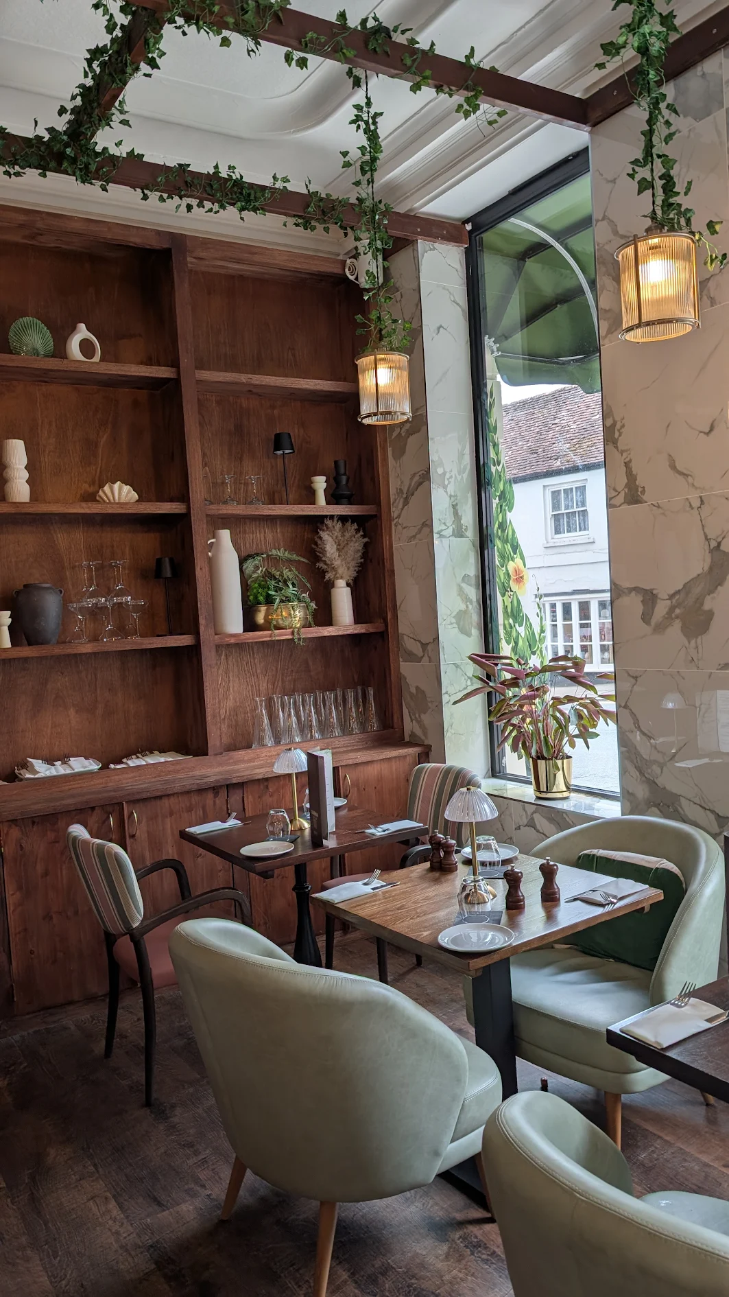 village taverna1 Wooden shelving filled with decorative vases and greenery stands beside dining tables paired with light green chairs.
