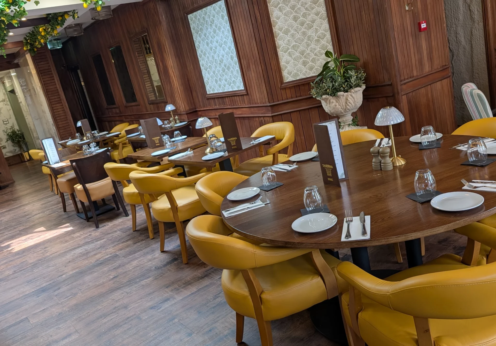 village taverna2 Round wooden tables with mustard-yellow chairs and Taverna menus fill a room featuring wood-paneled walls.