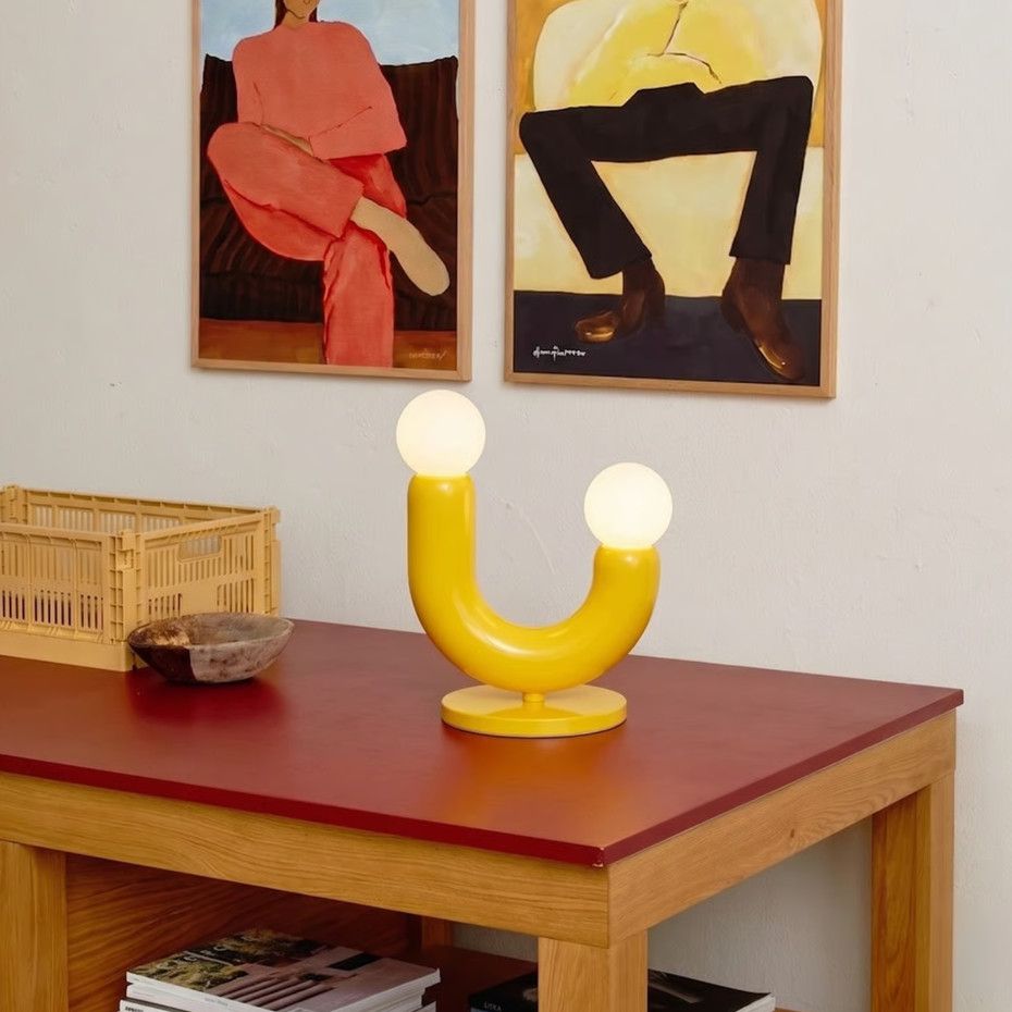 Play II table lamp with a vibrant yellow U-shaped body and dual spherical globes resting on a red table near wall art.