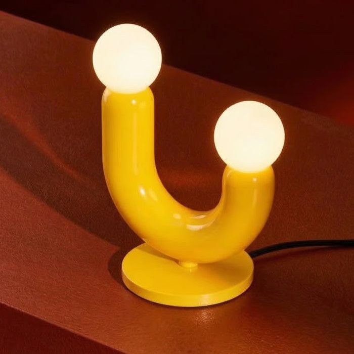 Play II table lamp featuring a curved yellow metal frame and two glowing opal glass spheres on a dark red surface.