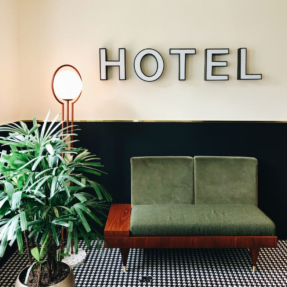 Frame floor lamp with warm bronze finish stands next to a green sofa in a hotel lobby with a large wall sign.