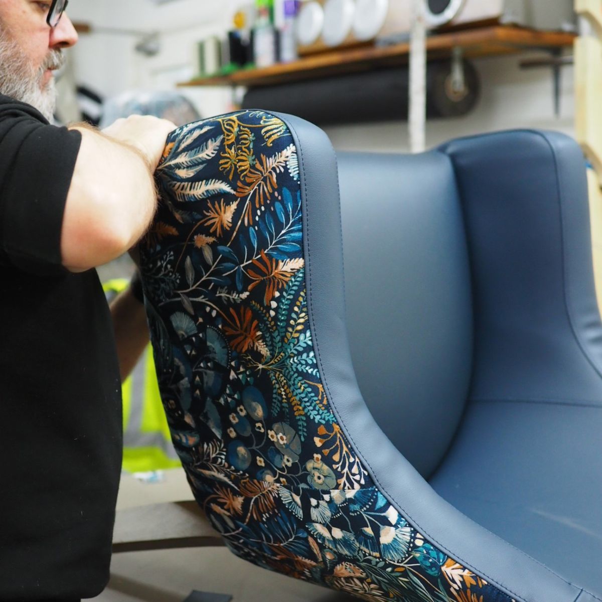 Upholsterer smooths navy blue botanical fabric onto the side panel of a chair featuring solid smooth leather accents.