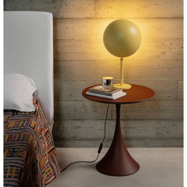 Leo coffee table featuring a deep red round top and pedestal base, positioned bedside with a round yellow lamp and books.