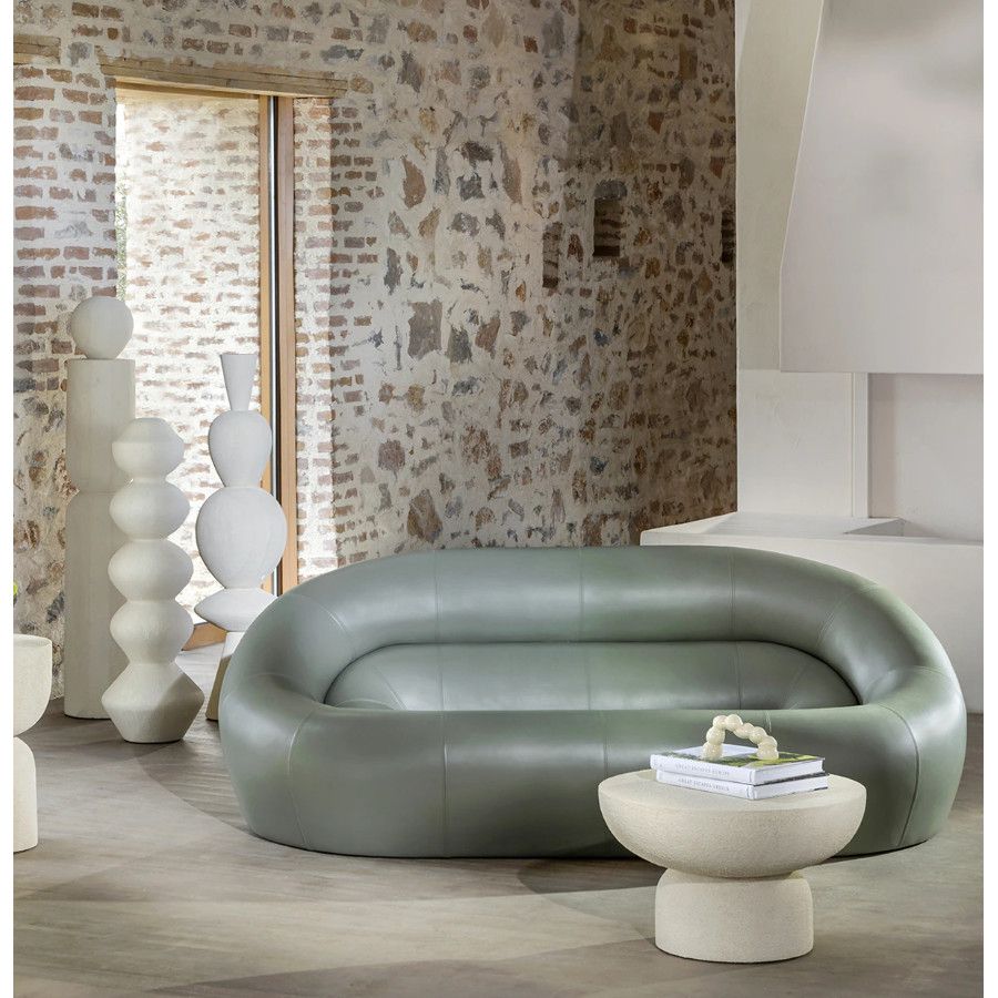 Front view of the Loop Sofa, featuring a full-width curved sage frame set against a textured rustic stone wall interior.