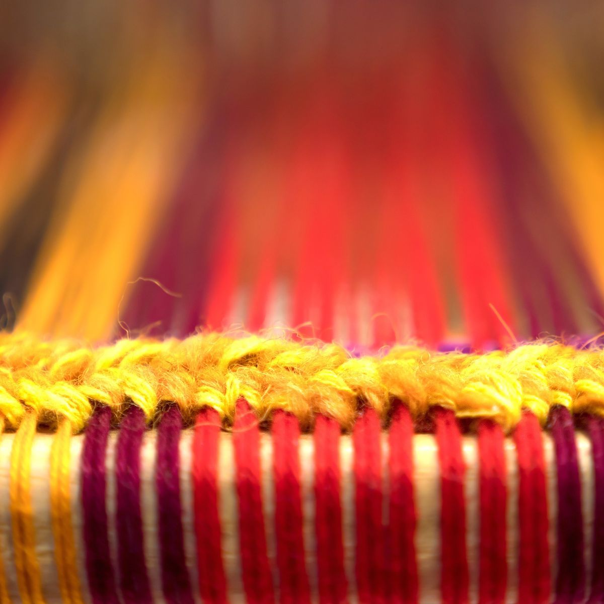 wools Fuzzy yellow wool yarn loops over taut red and purple vertical threads on a wooden loom.