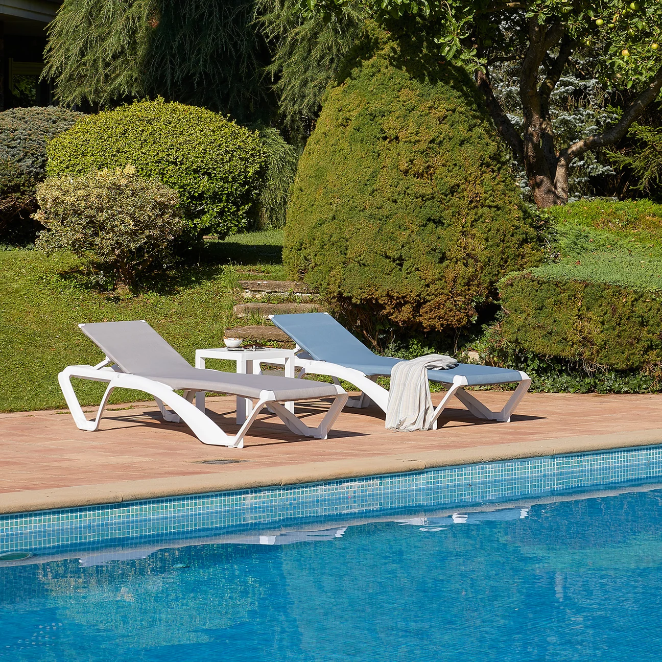Close-up of Mimi sun loungers with white frames and grey fabric, positioned on a patio beside a swimming pool.