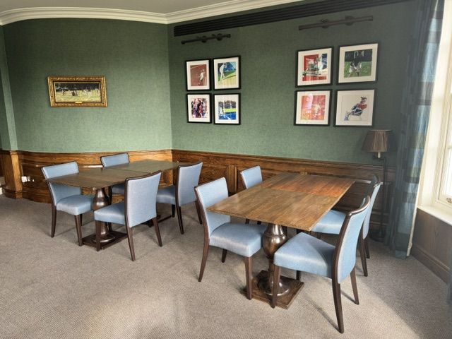 Two square wooden tables with turned pedestal bases surrounded by blue upholstered chairs in a green-walled room.