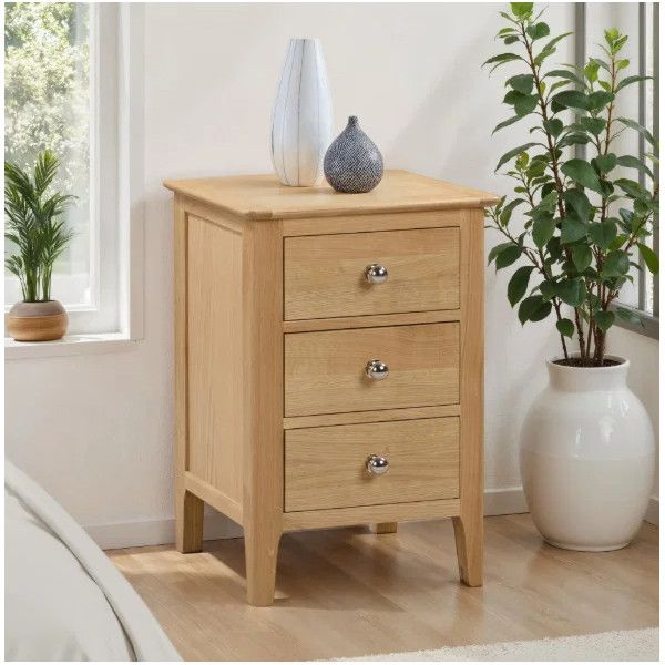 Cotswold Bedside table in light oak finish, showcasing a three-drawer design with metallic knobs in a sunlit room.