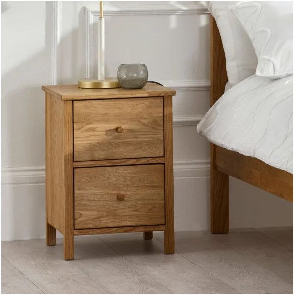 Natural oak Coxmoor 2 draw bedside placed beside a bed, topped with a minimalist table lamp and small glass bowl.