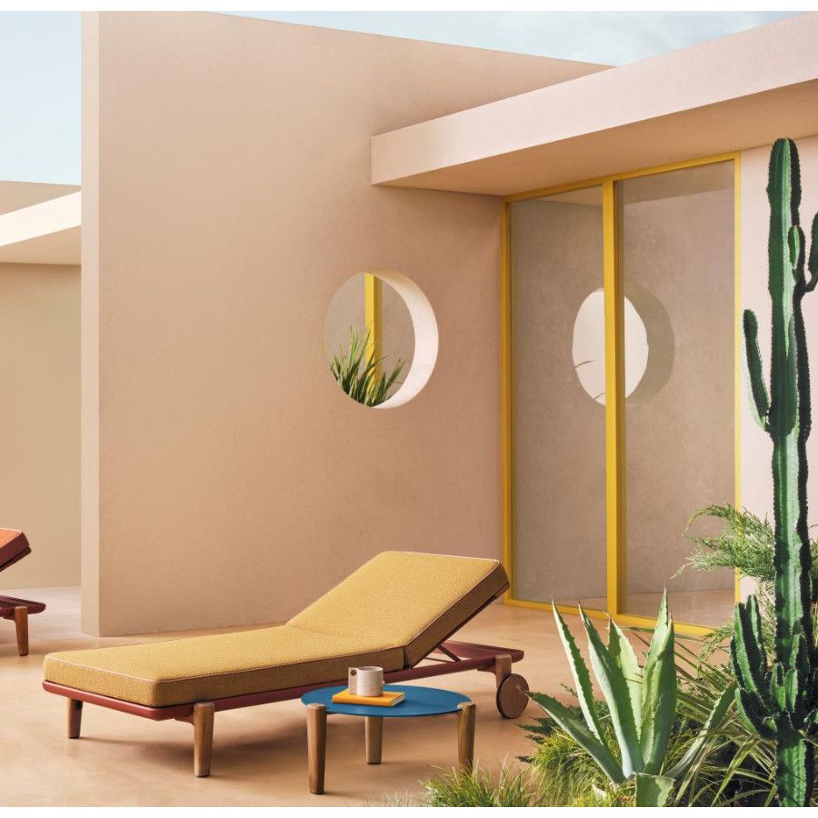 Fibra W Sun Lounger featuring a mustard cushion and reddish frame, positioned outdoors near a round table and cacti.