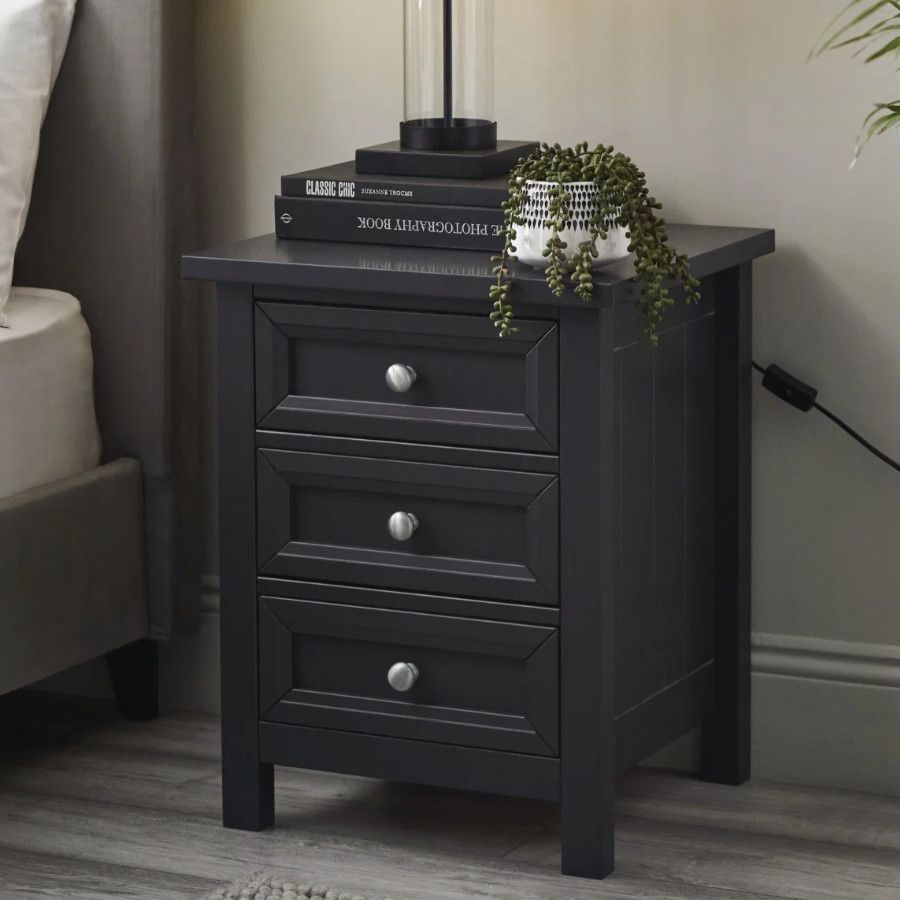 Maine bedside table in charcoal grey with three drawers, styled with books and a small plant in a bedroom setting.