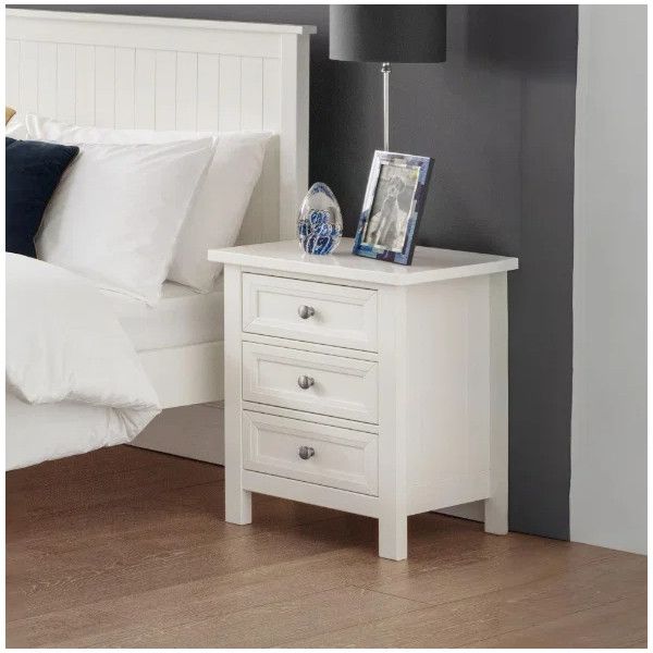 White Maine bedside table with three drawers, positioned next to a bed with a lamp and decorative picture frame.